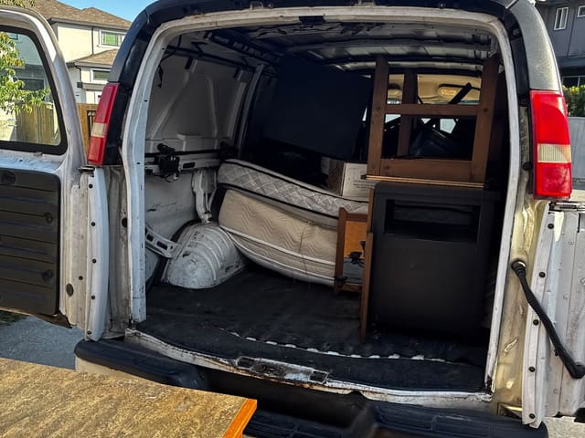 Open rear doors of a white van loaded with mattresses, wooden chairs and a dresser